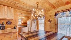 Interior de una cabaña de madera con una gran mesa de comedor, un candelabro y una zona de cocina.