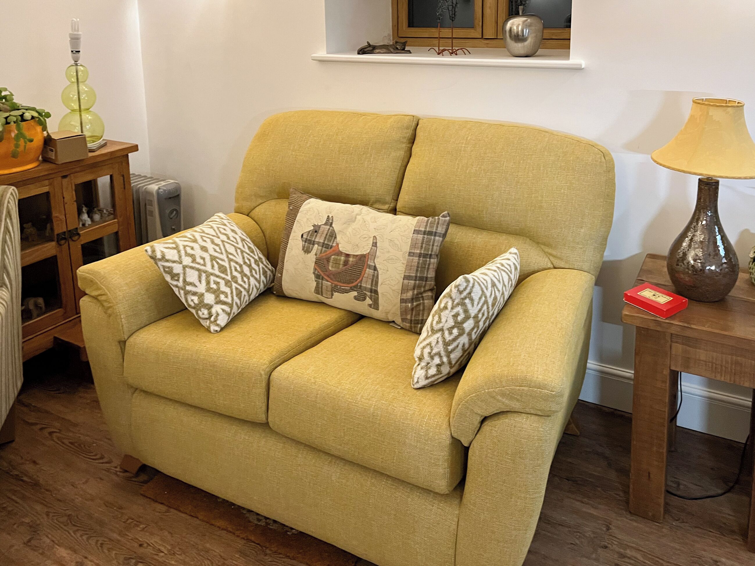 A yellow sofa with patterned cushions, a wooden side table with a lamp, and a cabinet with decorative items.