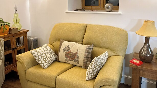Un sofá amarillo con cojines estampados, una mesita de madera con una lámpara y un mueble con objetos decorativos.
