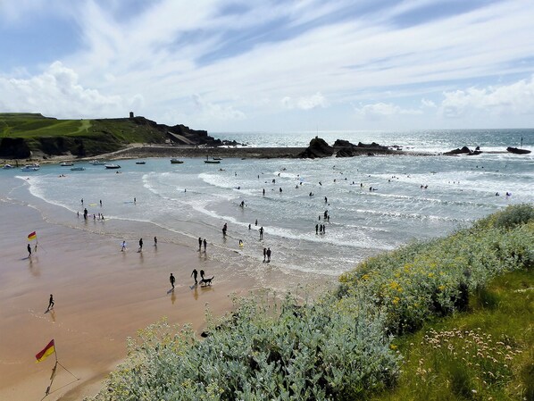 A beach with people walking and standing in the water, boats in the background, and a flag on the sand.