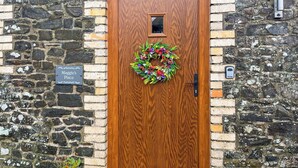 Una puerta de madera con un adorno de guirnalda, un muro de piedra, una planta en maceta y un letrero que dice 'Maggie's Place'.