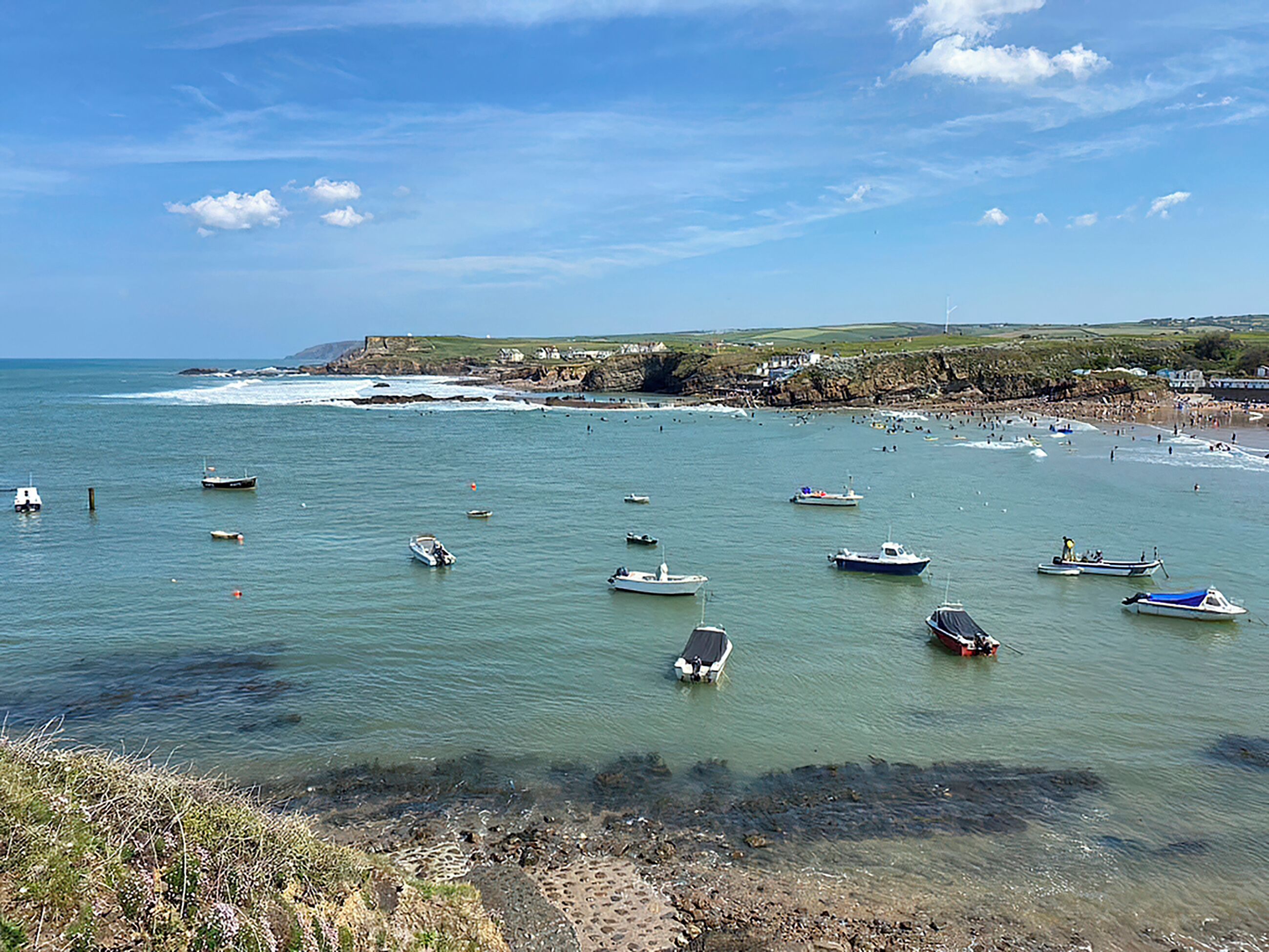 Uma paisagem costeira com barcos em águas calmas, uma praia de areia e um litoral rochoso.