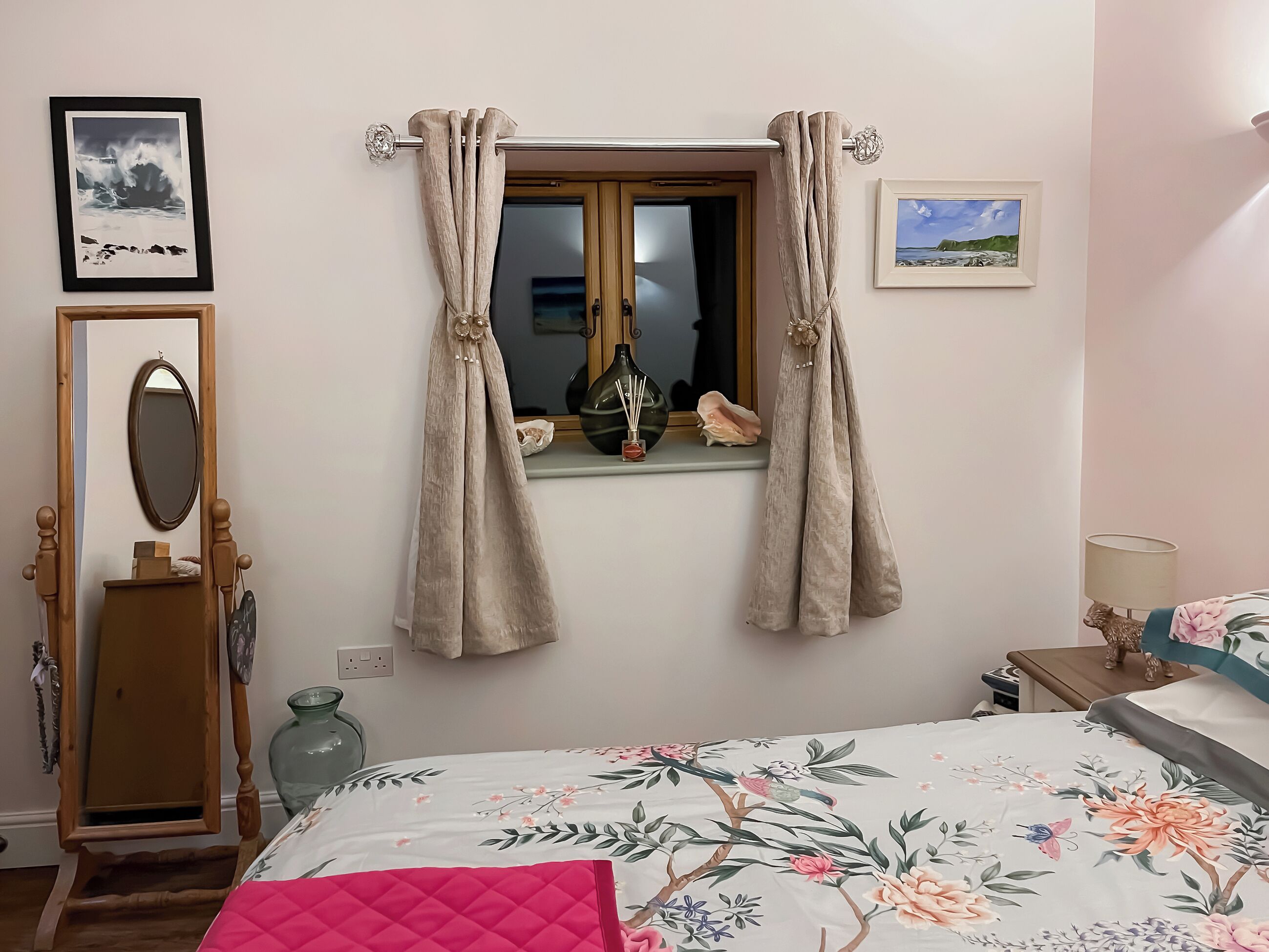 A bedroom with a bed, a window with curtains, a mirror, and a wall with two framed pictures.