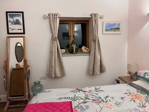 Un dormitorio con cama, ventana con cortinas, espejo y dos cuadros en la pared.