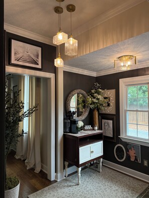 A room with a dark wall, a white-framed picture, a desk with a mirror, a vase with flowers, and a window with a view. - Masters Rental - Augusta, Ga. 1/2 mile from Augusta National (Augusta)