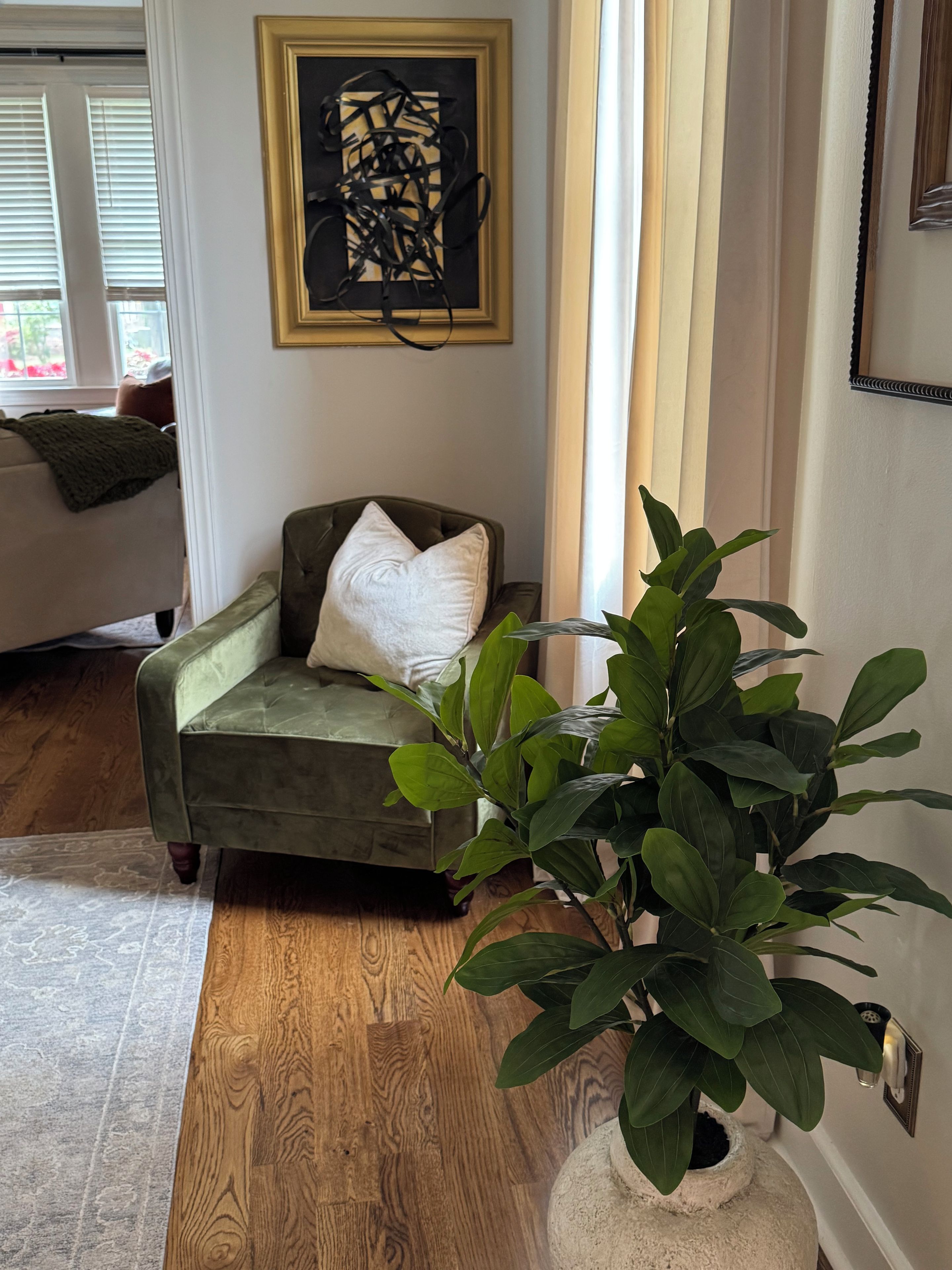 Una habitación con un sofá verde, una almohada blanca, una planta verde grande en una maceta blanca y un cuadro abstracto enmarcado en la pared.