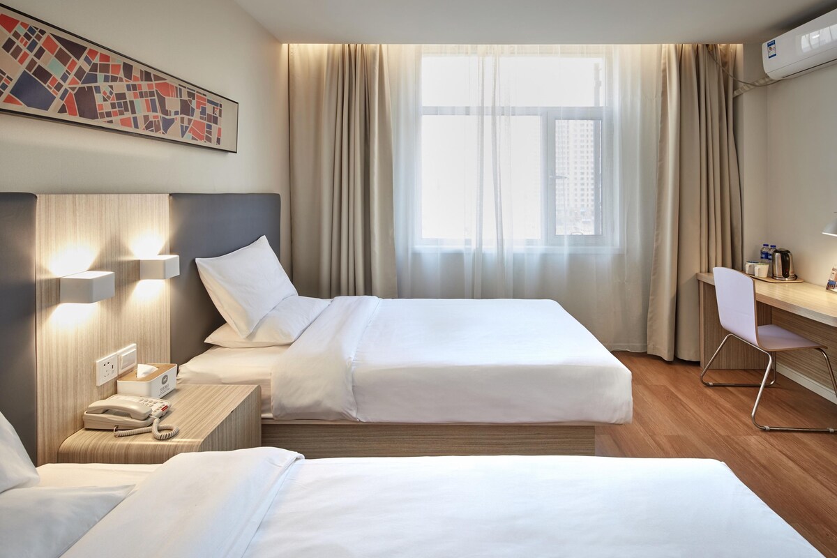Quarto de hotel com duas camas, uma escrivaninha, uma cadeira, telefone, chaleira e janela com cortinas.