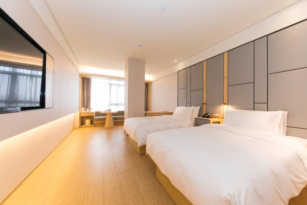 A modern hotel room with two beds, a sofa, a small table, and a large window.