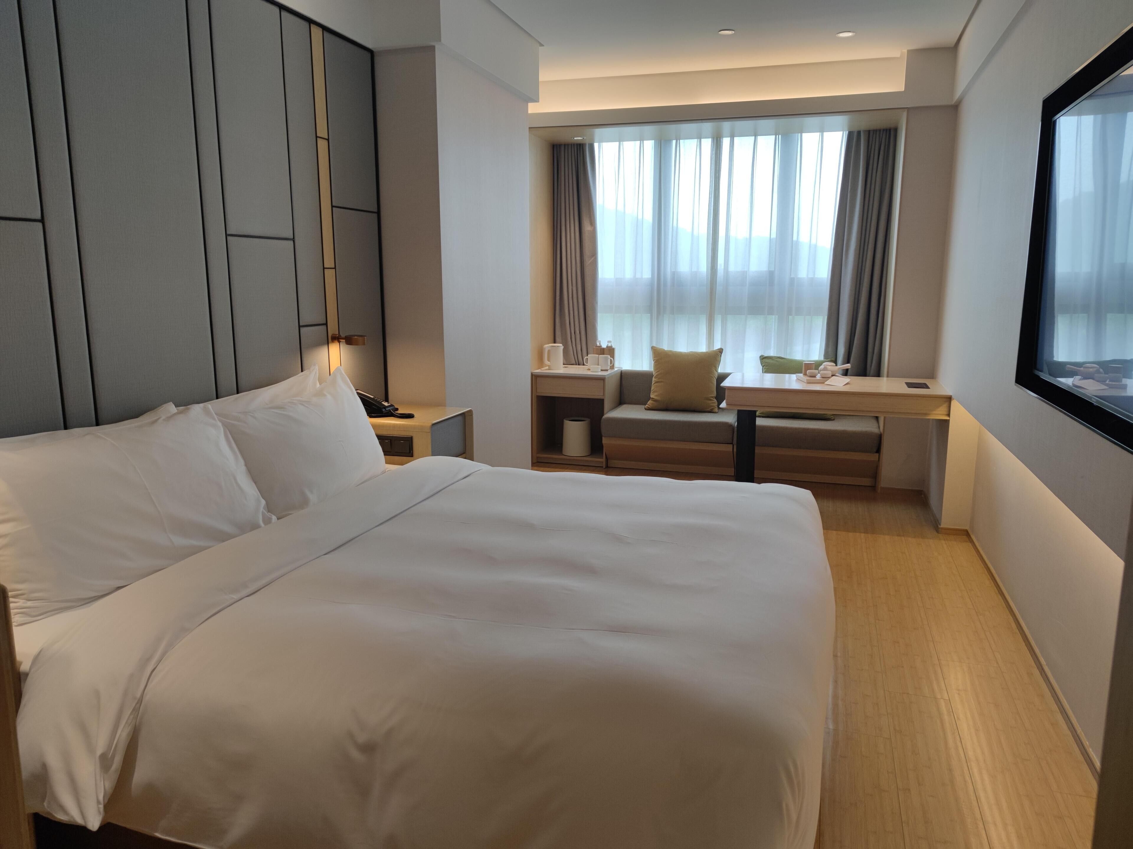 Una habitación de hotel con una cama grande, un escritorio con silla, una ventana con cortinas y una mesita de noche con lámpara.