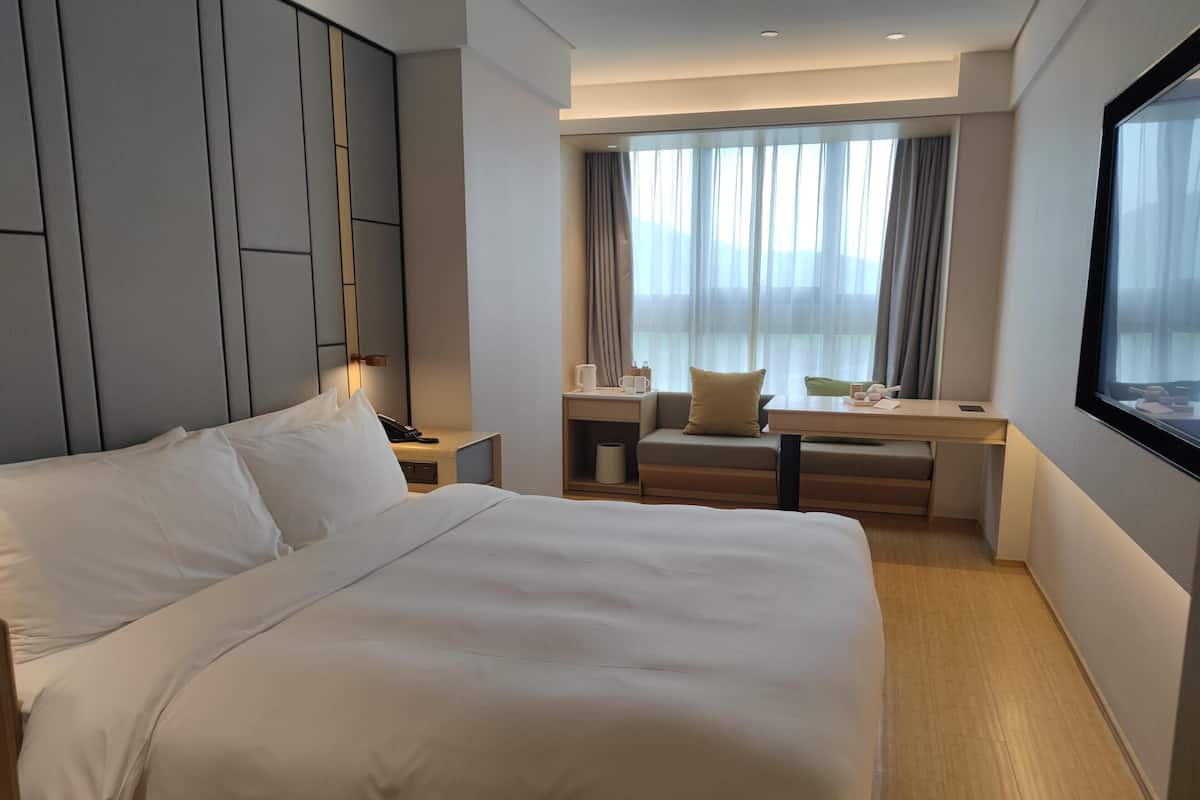 Una habitación de hotel con una cama grande, un escritorio con silla, una ventana con cortinas y una mesita de noche con lámpara.