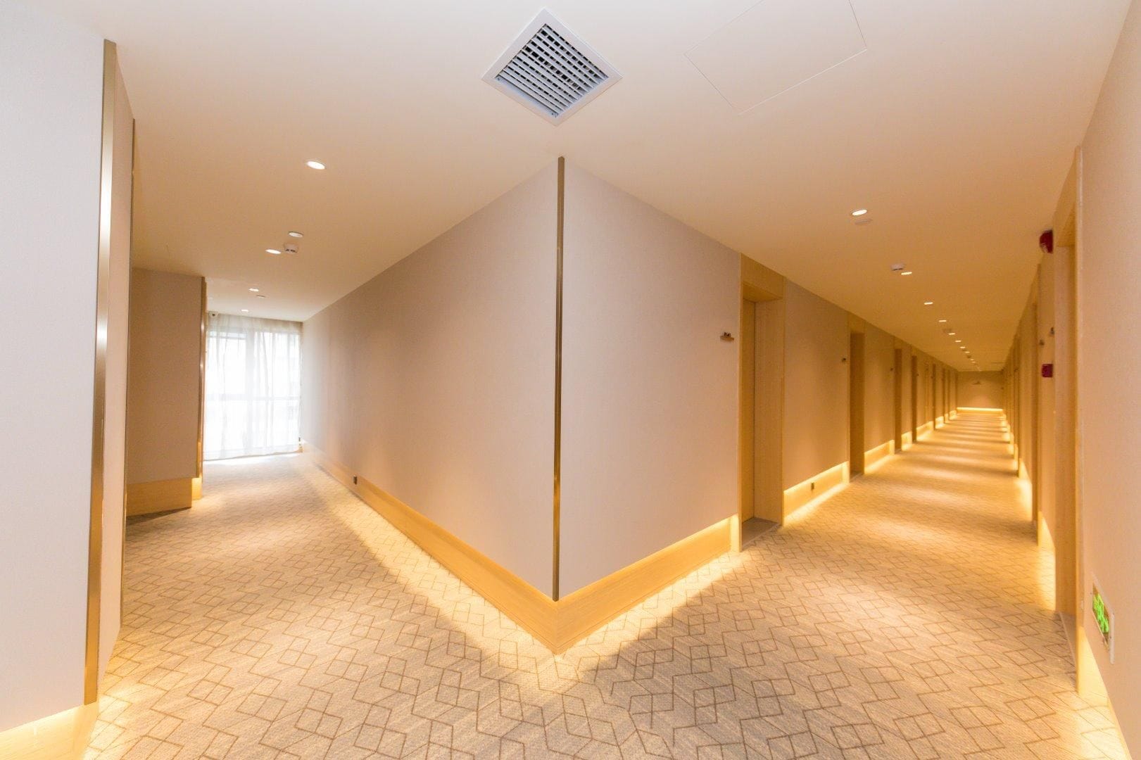 Un largo pasillo bien iluminado, con suelo estampado, una lámpara central y una rejilla de ventilación en el techo.