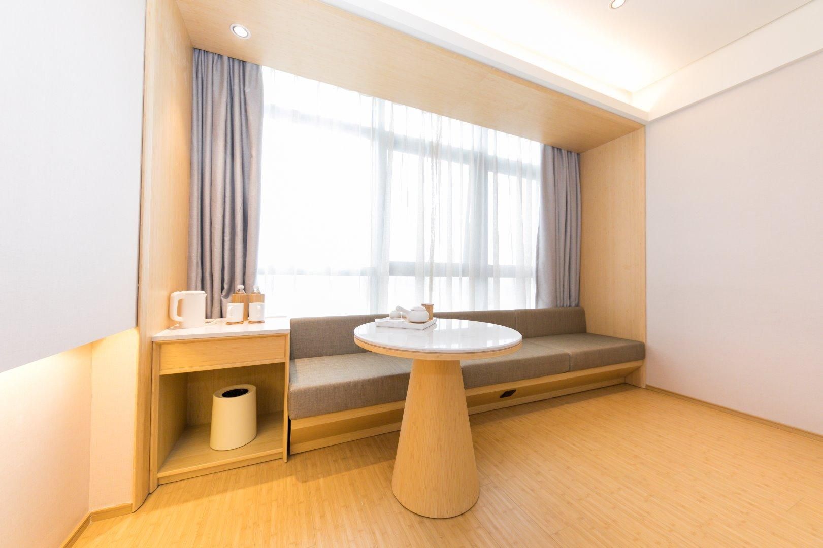 Habitació d'hotel moderna amb una taula rodona de fusta, una cadira amb coixí i una finestra gran amb cortines de tela fina.