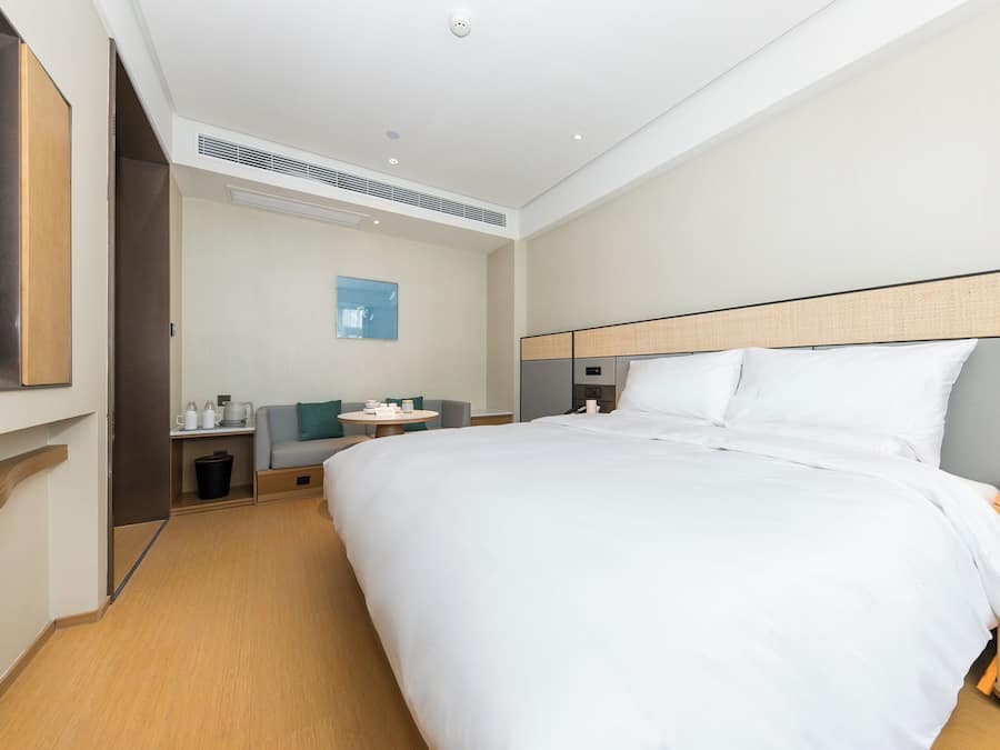 A modern hotel room with a large bed, a TV mounted on the wall, a small table with a chair, and a waste bin.