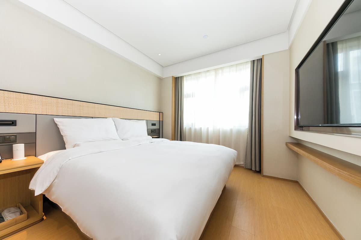 Una habitación de hotel con una cama grande, una mesita de noche con lámpara, un televisor montado en la pared y una ventana con cortinas.