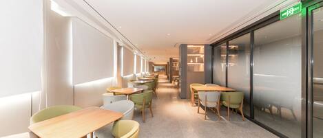 A corridor with tables and chairs, a green exit sign, and a glass wall on the right.