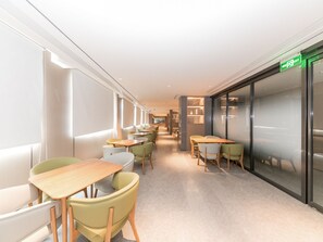 Un couloir avec des tables et des chaises, un panneau de signalisation verte indiquant la sortie, et un mur de verre sur la droite.