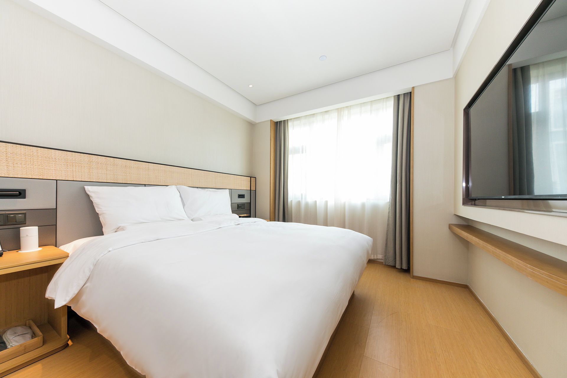 A hotel room with a large bed, a bedside table with a lamp, a TV mounted on the wall, and a window with curtains.