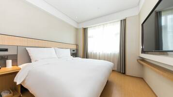 A hotel room with a large bed, a bedside table with a lamp, a TV mounted on the wall, and a window with curtains.