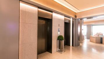 A modern elevator area with a clean, minimalist design, featuring a central planter with a small tree, and a reception desk in the background.