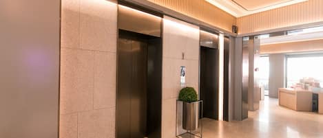 A modern elevator area with a clean, minimalist design, featuring a central planter with a small tree, and a reception desk in the background.