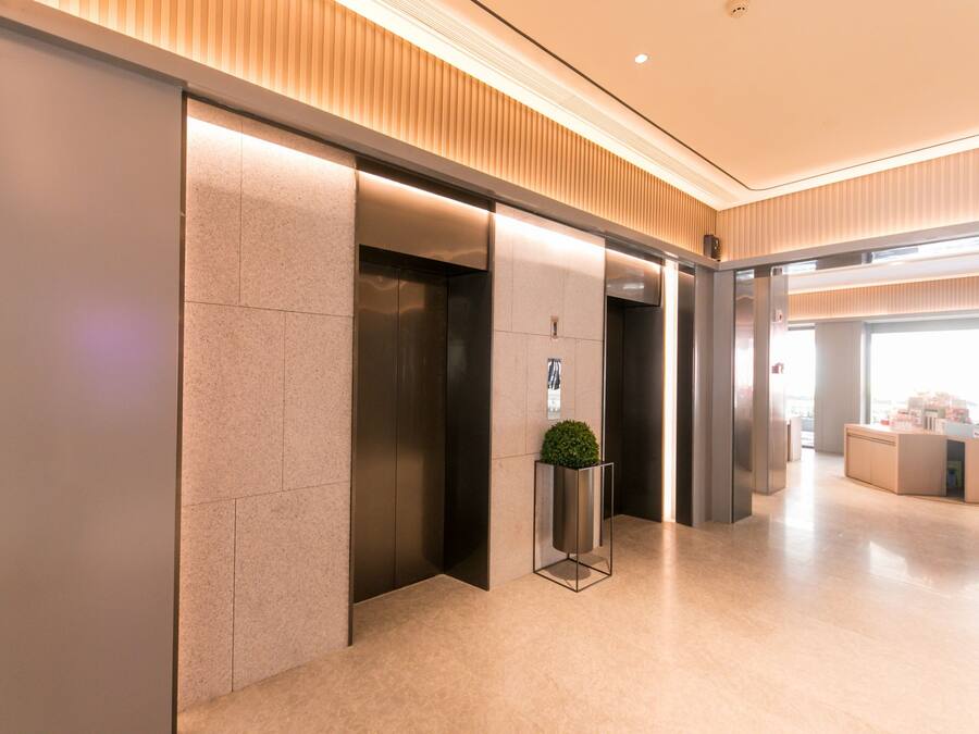 A modern elevator area with a clean, minimalist design, featuring a central planter with a small tree, and a reception desk in the background.