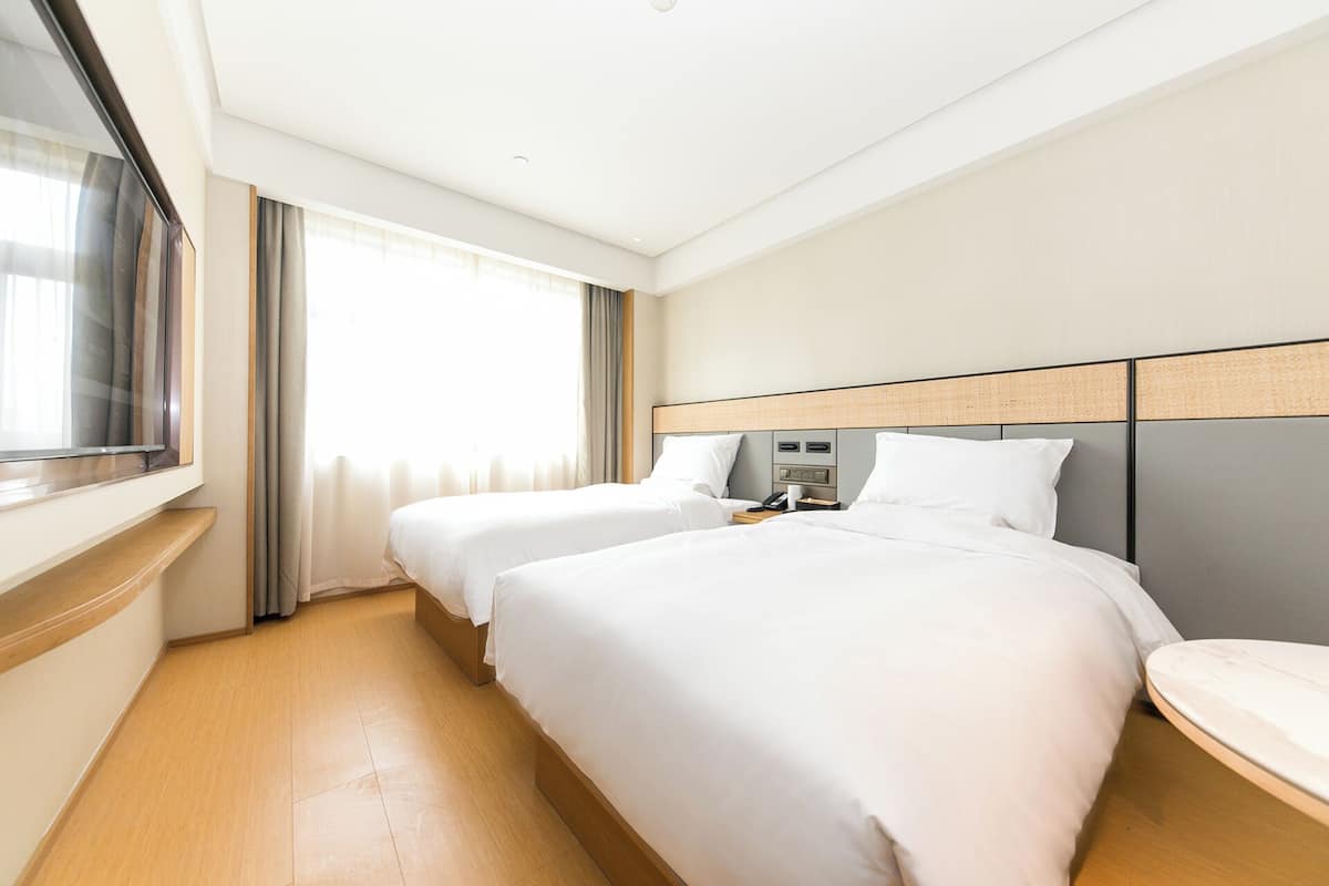 Habitación de hotel con dos camas, una mesita de noche con teléfono y lámpara, una ventana con cortinas y vistas al exterior.