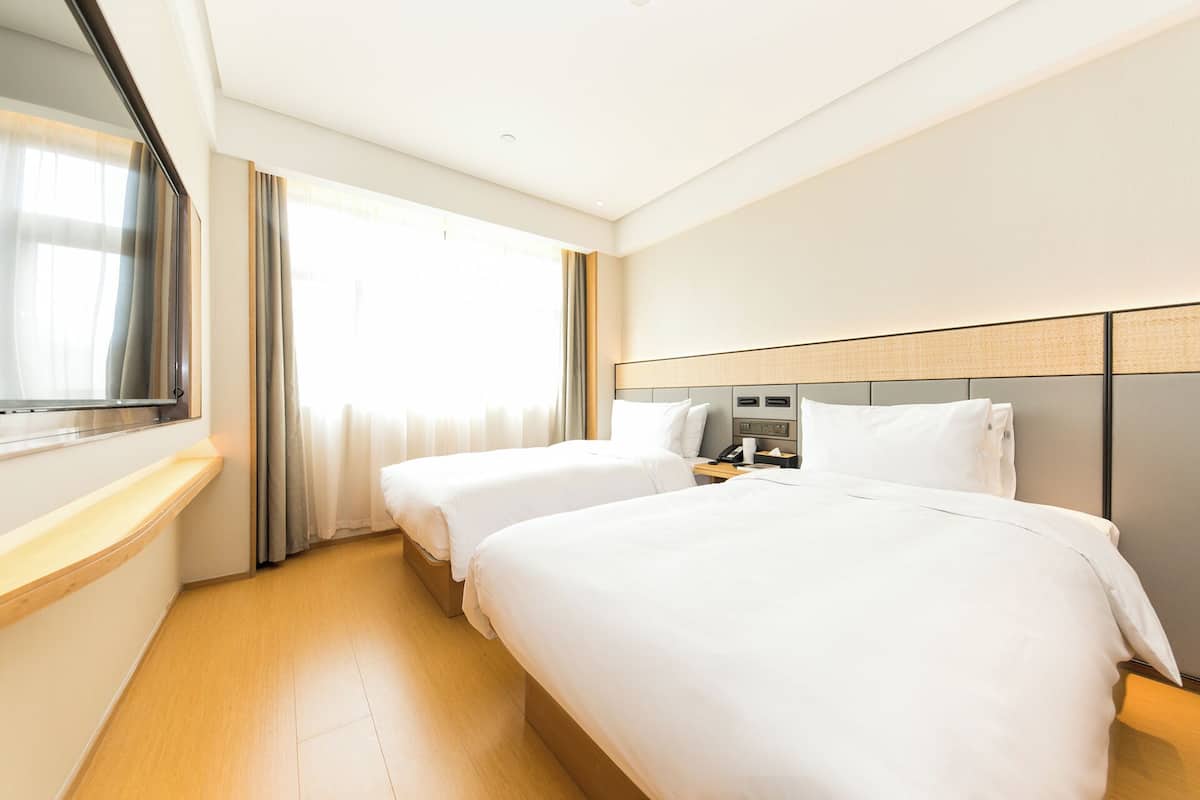 Habitación de hotel con dos camas, una mesita de noche con un teléfono y una taza, un ventanal con cortinas y un televisor de pantalla plana montado en la pared.