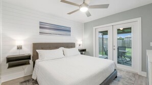 A bedroom with a bed, a ceiling fan, a nightstand, a wall-mounted lamp, and a sliding glass door leading to an outdoor patio. - Completely Remodeled Beach Getaway – Walk to Beach (Panama City Beach)