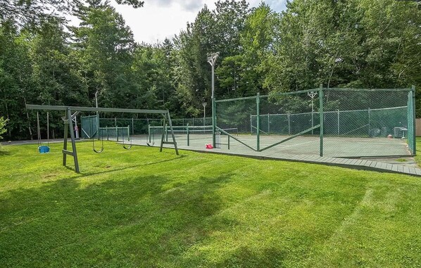 A fenced tennis court with a swing set and a basketball hoop in a park.