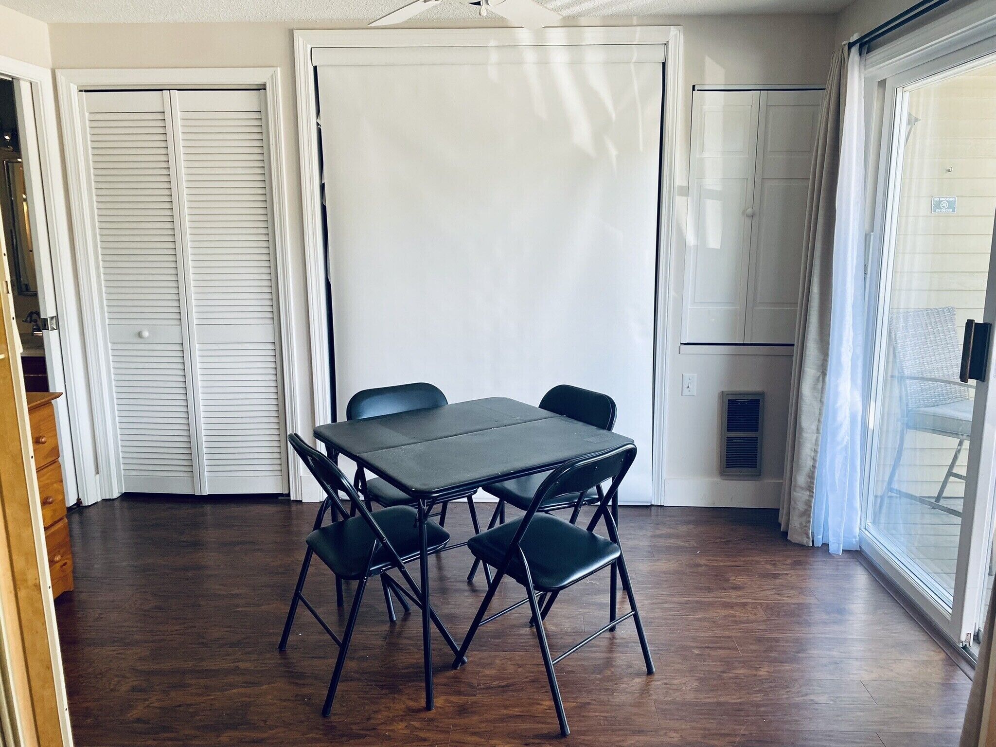 A room with a dark wooden floor, a table with four chairs, and a sliding glass door leading to a balcony.