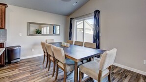 Un comedor con una mesa rectangular, seis sillas, un bote de basura, una encimera de cocina y un ventanal con cortinas.