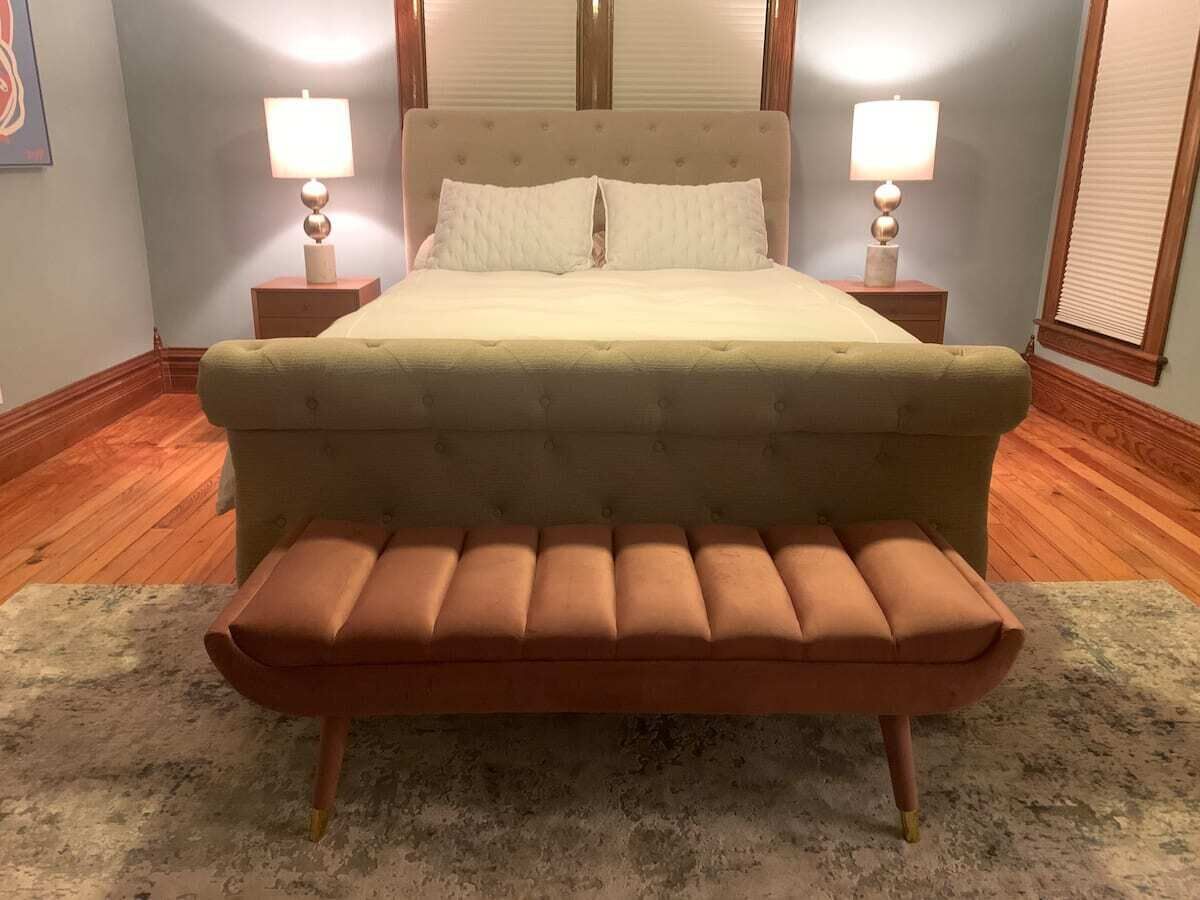 Una cama con cabecera capitonada, dos lámparas de noche y un banco frente a ella.