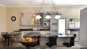 A modern kitchen with a central island, a microwave, an oven, and a sink. There are bar stools and a dining table with a fruit bowl. - Grande Plaisir Holiday Home (Franschhoek)