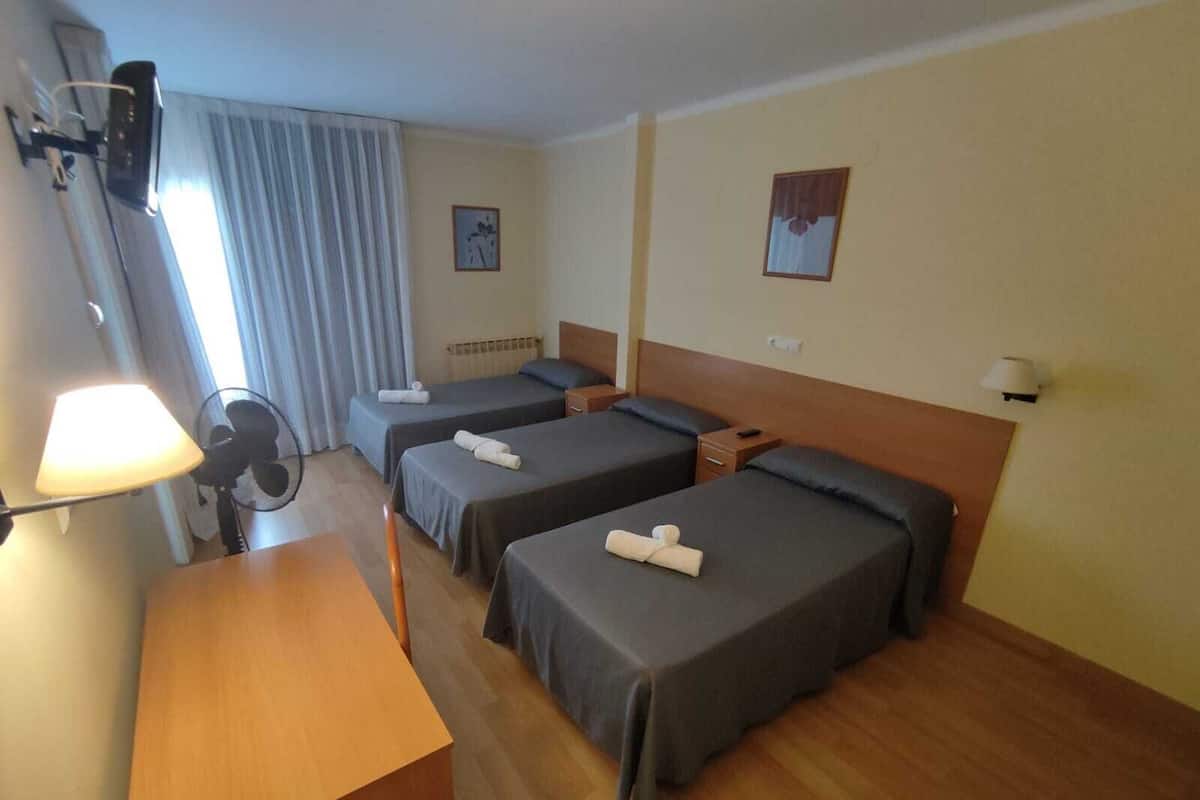 A hotel room with two single beds, a wooden desk, a chair, a desk lamp, a fan, and a television mounted on the wall.