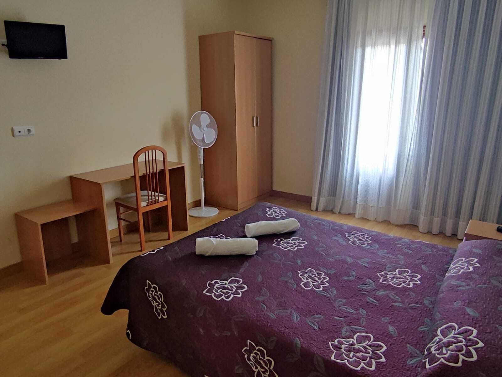 A bedroom with a bed, a desk, a chair, a wardrobe, a fan, and a TV on the wall.