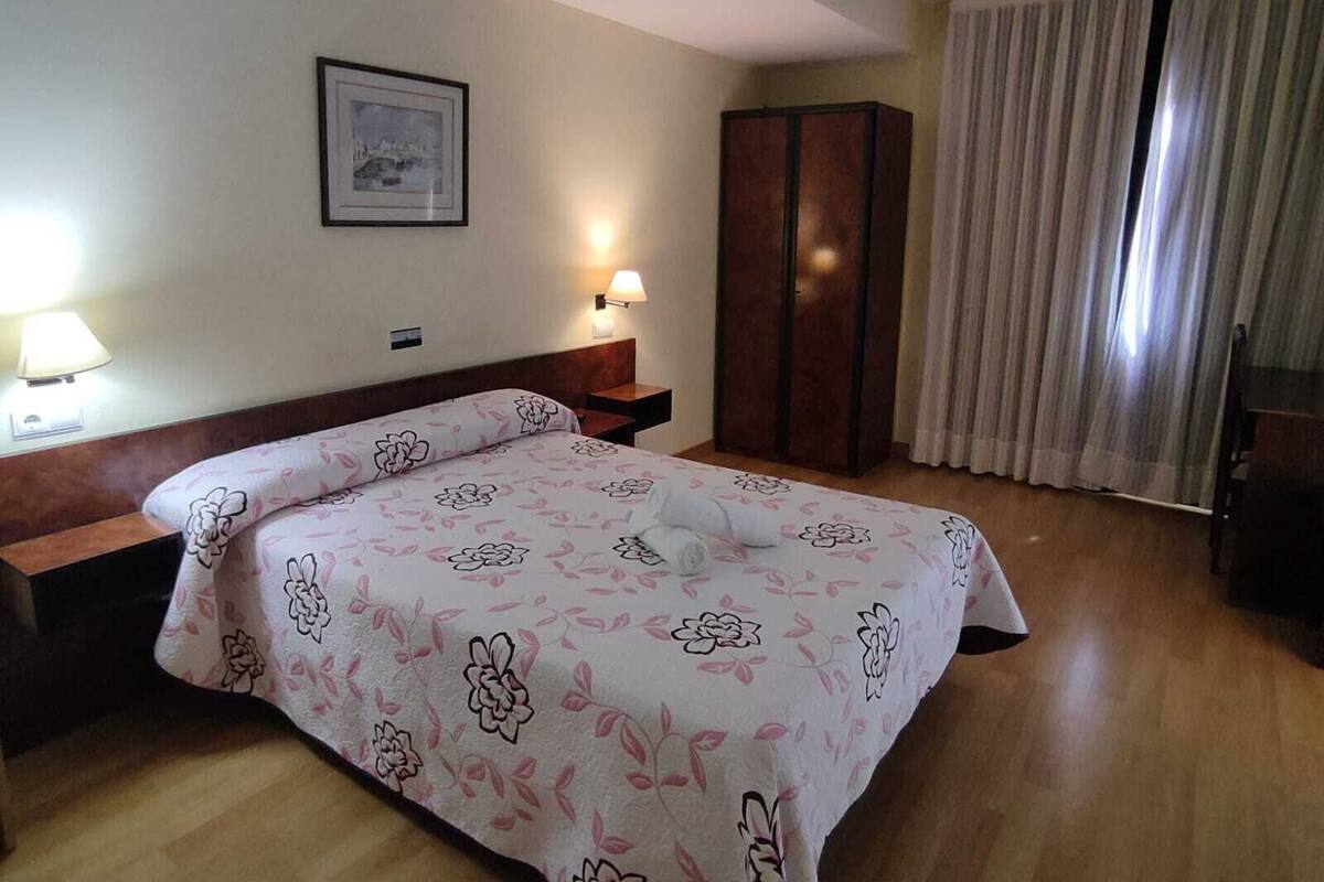 A hotel room with a bed, bedside tables, a wardrobe, a framed picture on the wall, and a window with curtains.