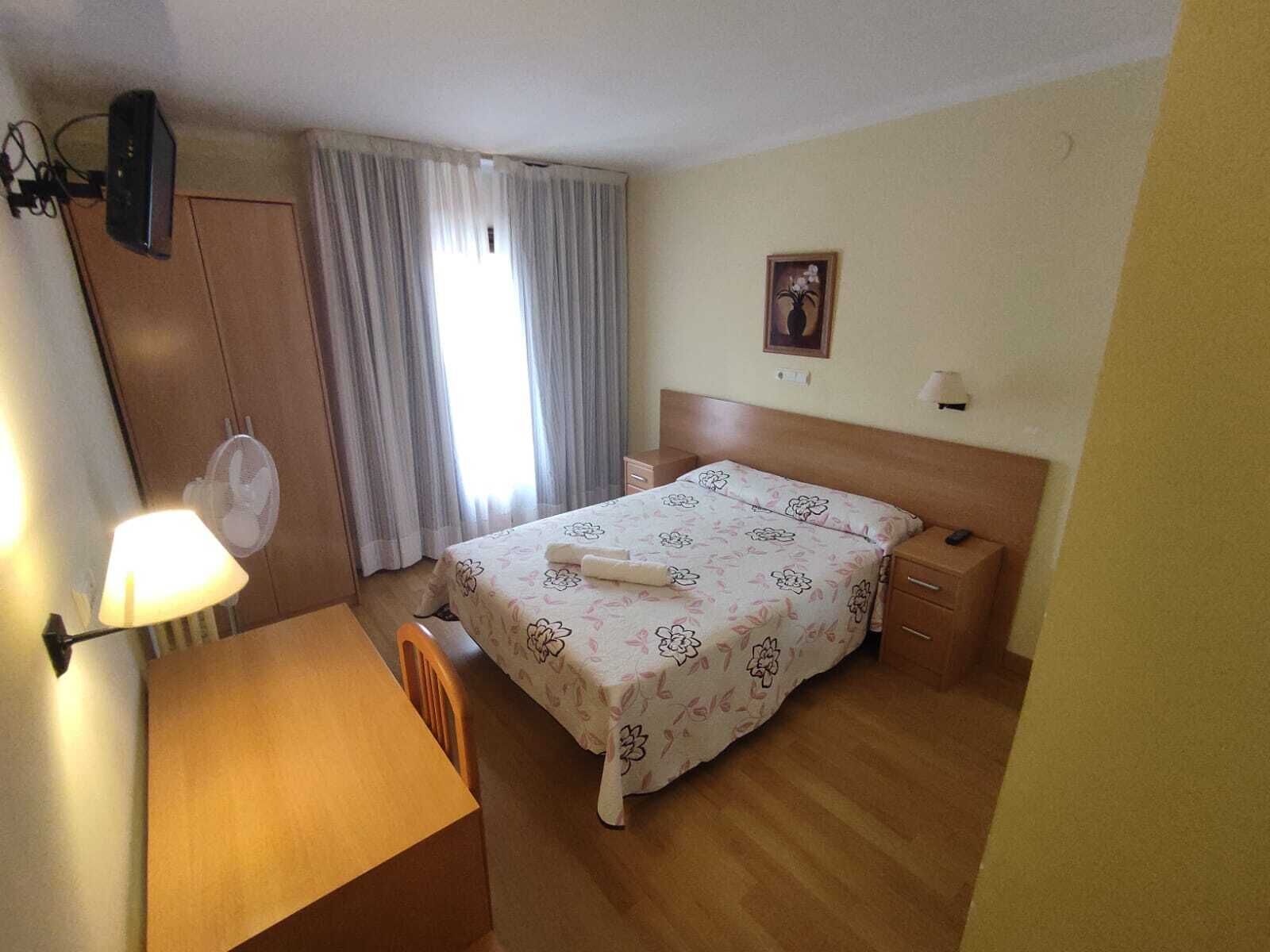 A hotel room with a bed, a desk, a chair, a nightstand, a lamp, a fan, and a window with curtains.
