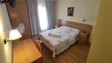A hotel room with a bed, a desk, a chair, a nightstand, a lamp, a fan, and a window with curtains.