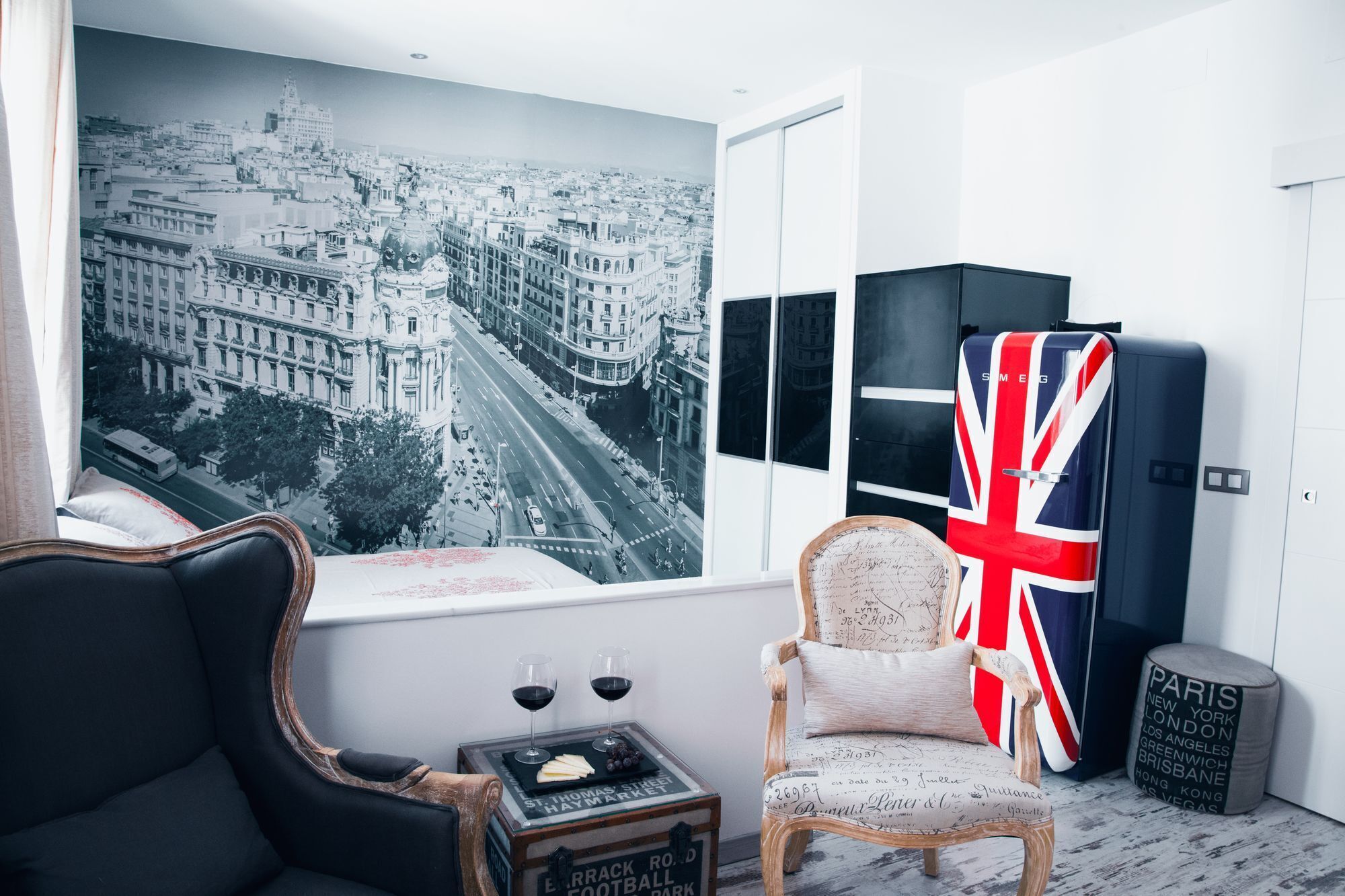 Ett rum med en stor väggmålning som föreställer en stadsbild, en fåtölj i vintage-stil, ett bord med glas och ost samt ett kylskåp med en Union Jack-design.