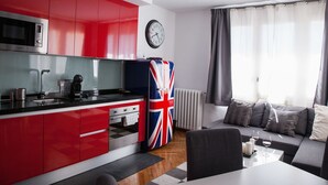 A modern kitchen with red cabinets, a microwave, and a refrigerator with a Union Jack flag. There is a dining area with a table and chairs, and a living room with a sofa and a window with curtains. - Suites Miguel Ángel - Adults Only (Madrid)