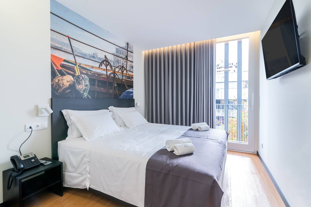 A hotel room with a bed, a nightstand with a phone, a painting on the wall, a window with curtains, and a TV mounted on the wall.