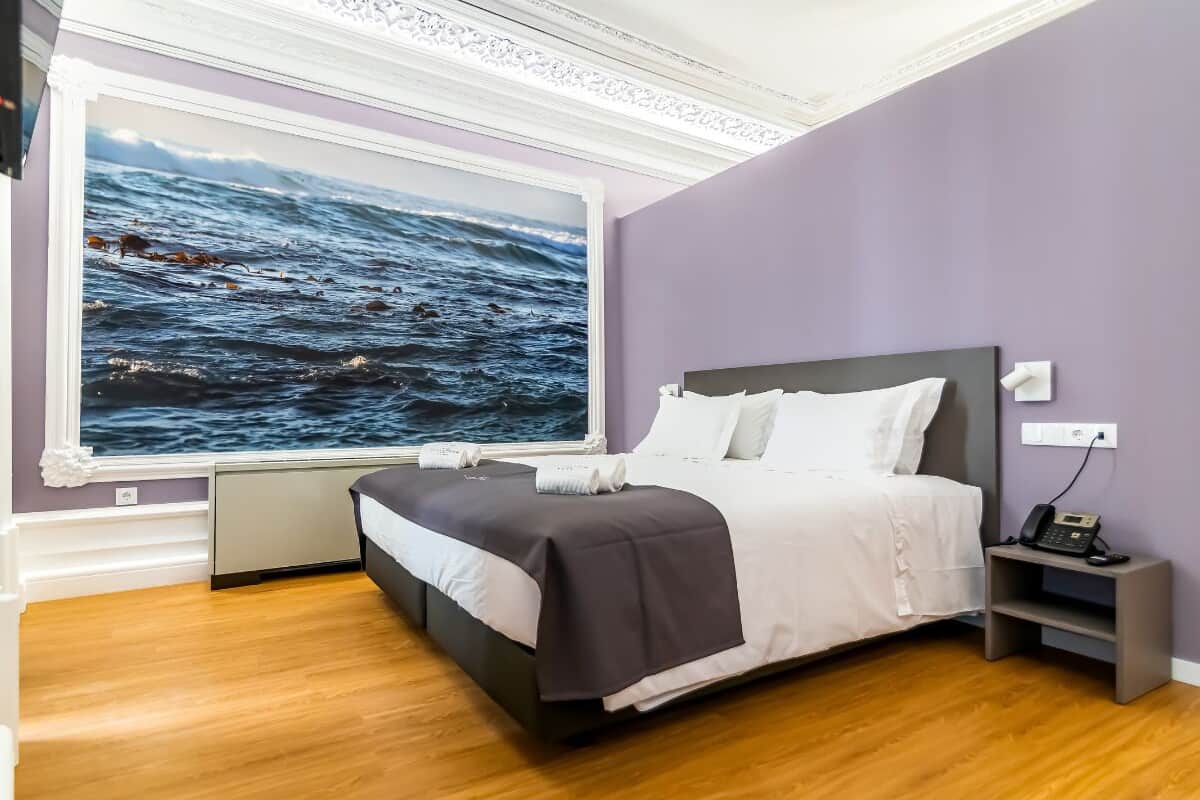 A modern bedroom with a large window, a bed with white and gray bedding, a bedside table with a phone, and a wall-mounted outlet.