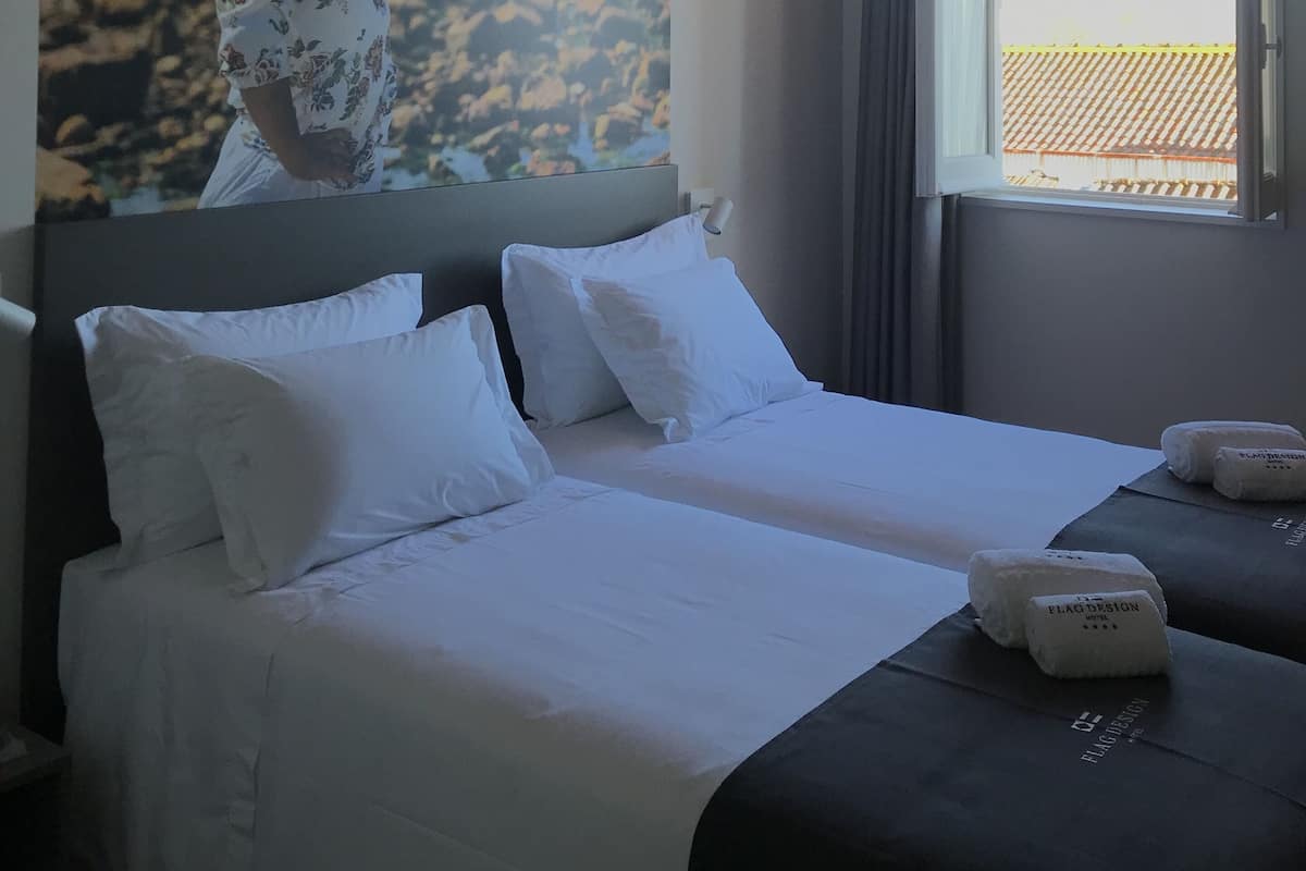 A hotel room with a bed, pillows, a nightstand, a window with curtains, and a wall mural.