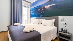 A hotel room with a bed, a painting of a coastal scene, a bedside table with a phone, and a window with curtains. - Flag Design Hotel (Viana do Castelo)