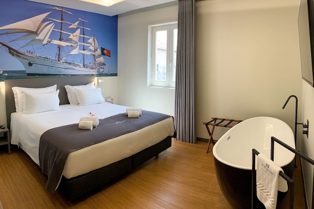 A hotel room with a large bed, a painting of a sailing ship, a bathtub, and a window with curtains.