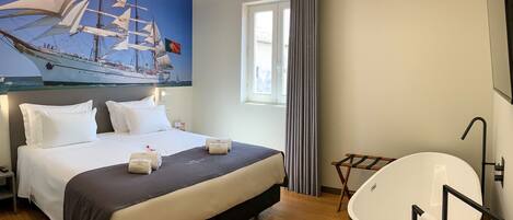 A hotel room with a large bed, a painting of a sailing ship, a bathtub, and a window with curtains.