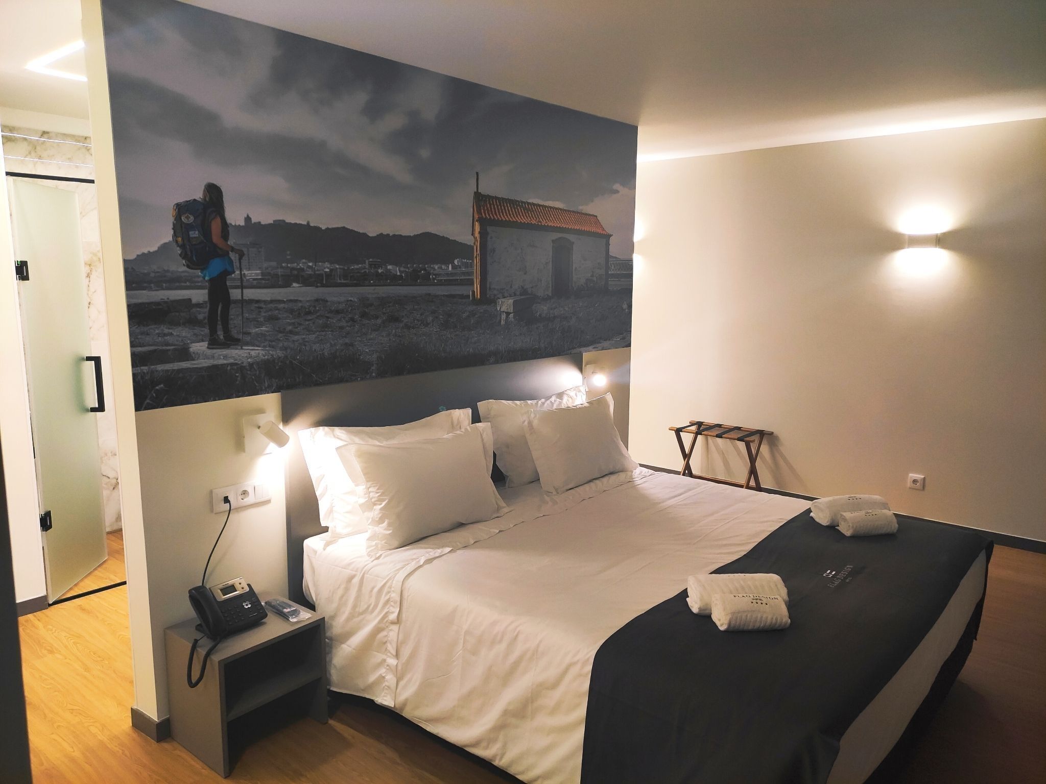 A hotel room with a bed, a nightstand with a phone, a wall-mounted light, and a painting of a person on a mountain.