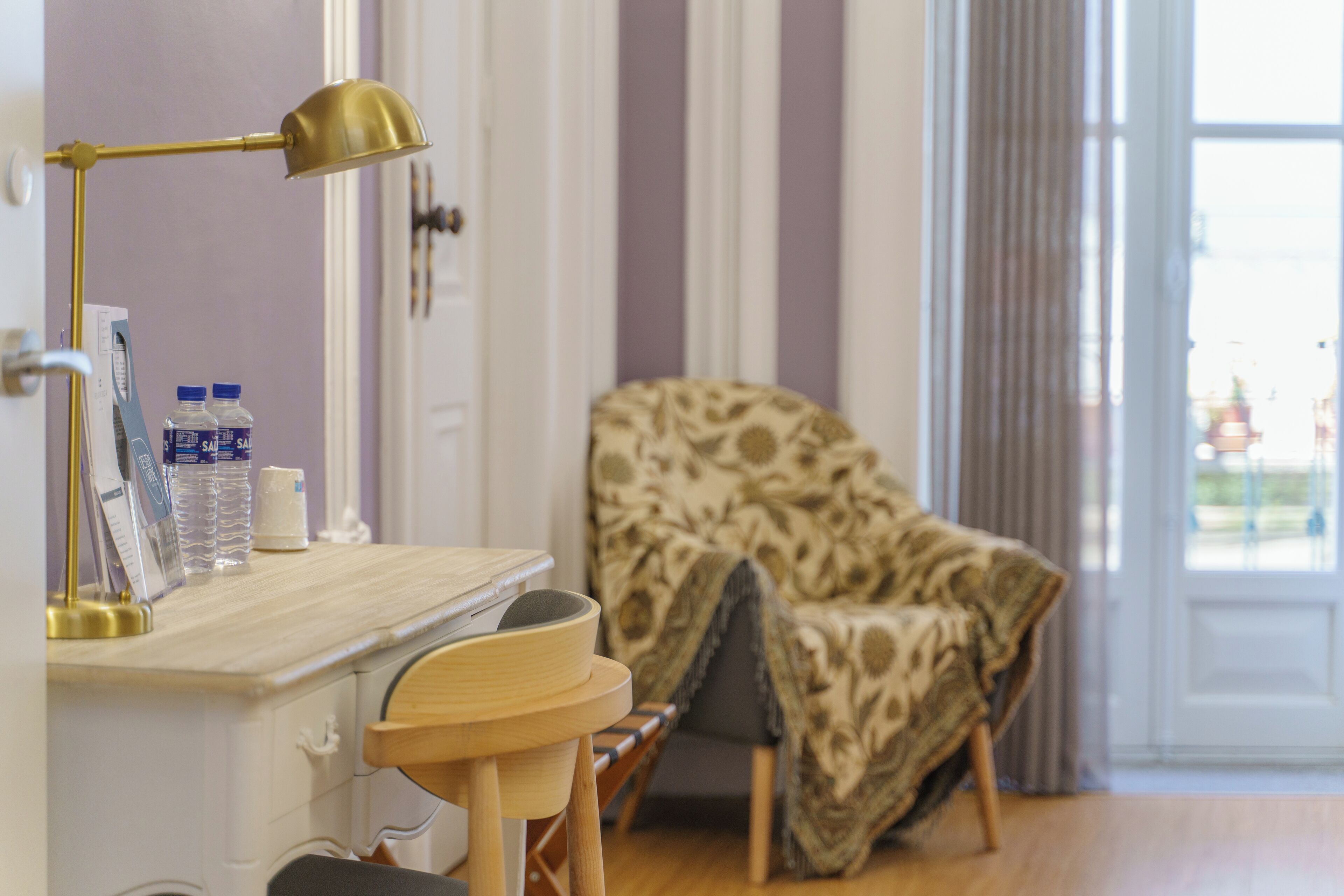 A room with a desk, a lamp, a chair, and a patterned armchair.