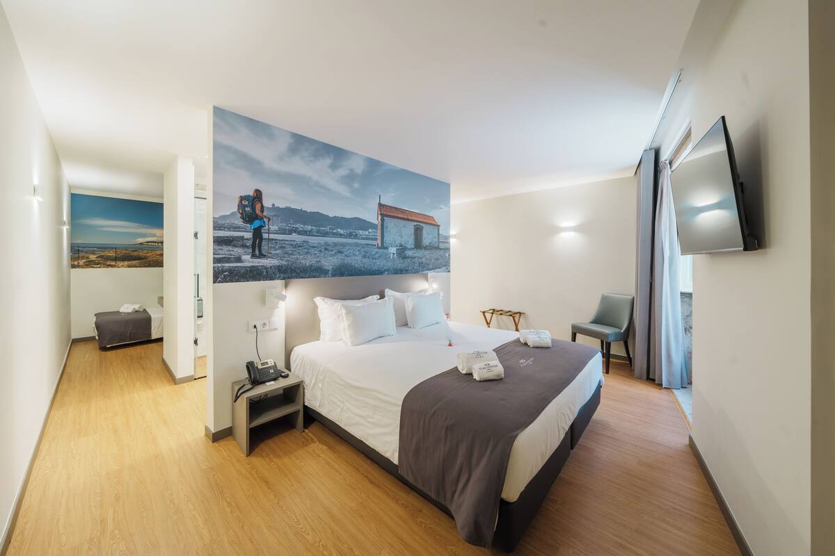 A modern hotel room with a large bed, a flat-screen TV, a desk with a lamp, and a painting of a hiker on a mountain.