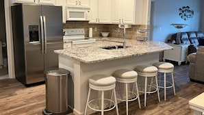 A modern kitchen with white cabinetry, a stainless steel refrigerator, a built-in microwave, and a granite countertop. There are three bar stools and a trash can.